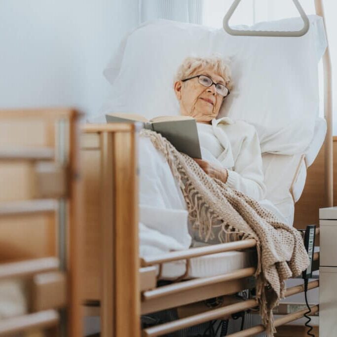 Senior Woman Lies In A Hospital Bed And Reads A Book